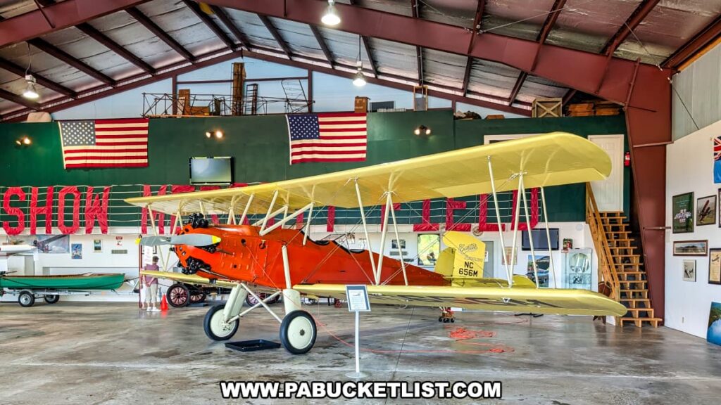 A yellow and orange vintage biplane displayed inside a hangar at the Eagles Mere Air and Auto Museum in Sullivan County, Pennsylvania, with American flags hanging on the green back wall, a staircase leading to a mezzanine level with additional aviation artifacts, and other classic aircraft and memorabilia visible throughout the spacious exhibit area.