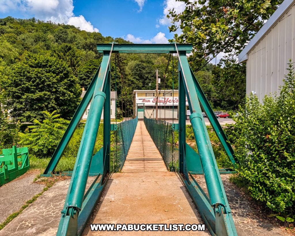 A view of the Hooversville Swinging Bridge in Somerset County, PA, showing the green metal supports and wooden walkway leading across the Stonycreek River, with trees, buildings, and a hillside in the background.