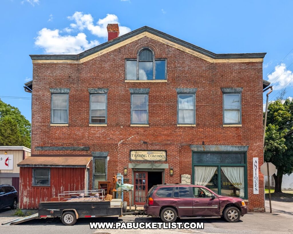 Exterior view of the Penn Avenue Trading Company in Newport, Perry County, PA, showing a large three-story red brick building with vintage-style windows, a faded wooden sign above the entrance, and an “Antiques” sign on the corner, with a maroon SUV and utility trailer parked in front under a bright blue sky with scattered clouds.