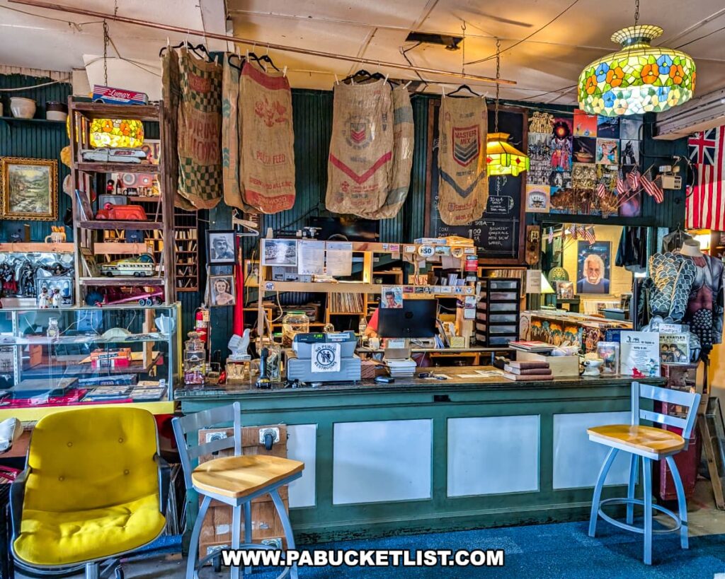 Front counter area of the Penn Avenue Trading Company in Newport, Perry County, PA, featuring vintage feed sacks hanging above the register, colorful stained glass pendant lights, a chalkboard menu, shelves filled with books and records, and displays of retro decor, collectibles, and small antiques, all creating a nostalgic general store atmosphere.