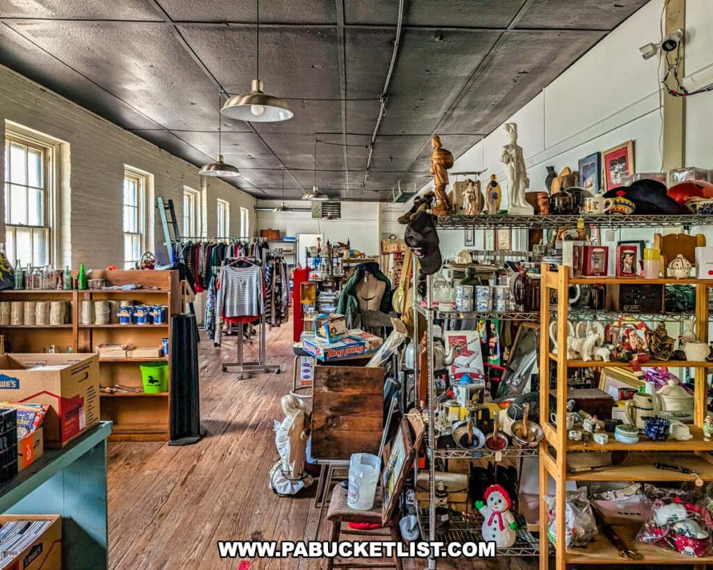 Second floor of the Penn Avenue Trading Company in Newport, Perry County, PA, featuring vendor booths filled with vintage clothing, books, toys, mugs, figurines, framed art, and assorted collectibles, all arranged in a bright space with wooden floors, exposed brick walls, large windows, and a black ceiling with hanging industrial lights.
