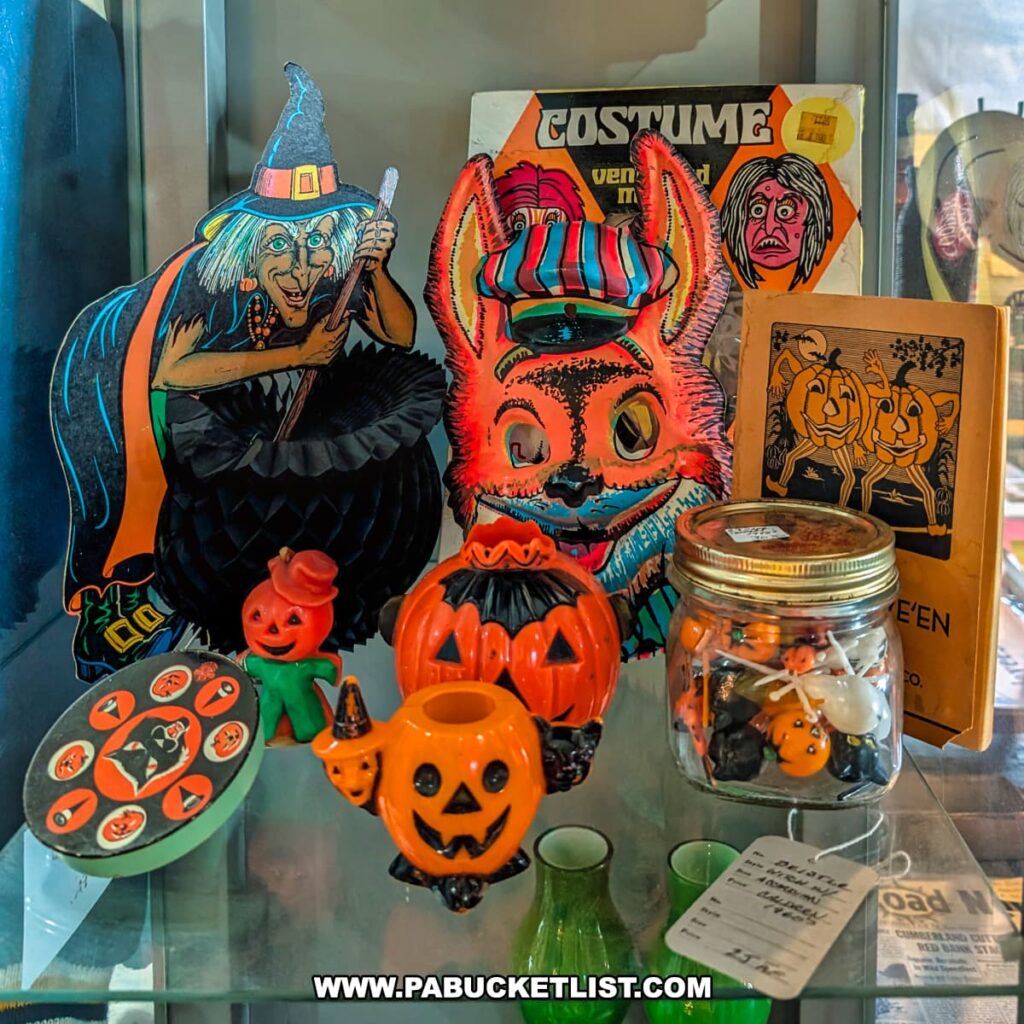 Display of vintage Halloween decorations at the Penn Avenue Trading Company in Newport, Perry County, PA, featuring colorful paper cutouts of a witch and a fox mask, ceramic pumpkin-themed figurines, a jar of small Halloween trinkets, an old costume box, and a booklet of classic Halloween imagery, all arranged in a glass case.