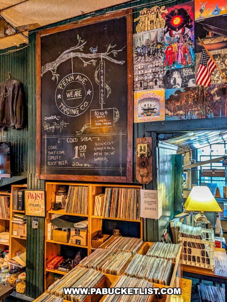 Vinyl record section at the Penn Avenue Trading Company in Newport, Perry County, PA, featuring crates and shelves filled with vintage albums beneath a large hand-drawn chalkboard sign listing store hours and cold soda prices, surrounded by colorful posters, retro memorabilia, and a cozy desk lamp that adds to the nostalgic atmosphere.