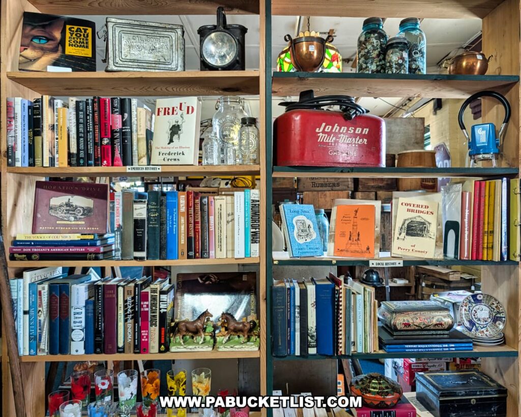 Bookshelf display at the Penn Avenue Trading Company in Newport, Perry County, PA, filled with vintage and local history books, glassware, mason jars of buttons, a red Johnson Mule-Master gas can, antique collectibles, and decorative items, all neatly arranged in a warm and inviting antique store setting.