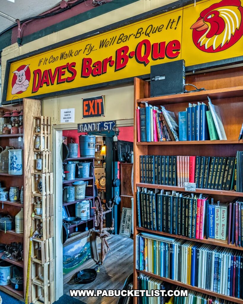 Interior corner of the Penn Avenue Trading Company in Newport, Perry County, PA, featuring shelves of vintage books, crocks, and collectibles beneath a large yellow "Dave’s Bar-B-Que" sign with the slogan "If it can walk or fly... we’ll bar-b-que it!", with a narrow hallway leading to more antique displays filled with stoneware, signs, and rustic décor.