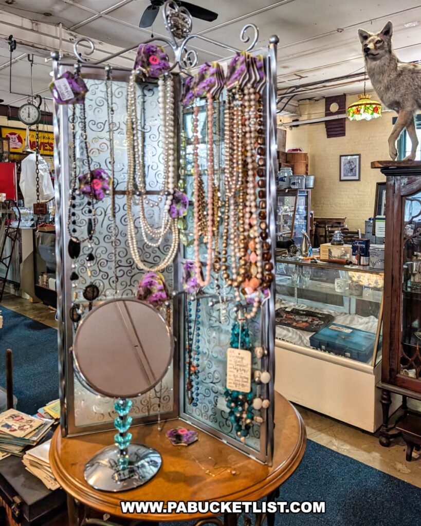Jewelry display at the Penn Avenue Trading Company in Newport, Perry County, PA, featuring a decorative metal screen adorned with vintage necklaces, beaded strands, costume jewelry, and a small mirror on a stand, with glass cases, antique furnishings, and a taxidermy coyote visible in the background of this eclectic multi-vendor antique shop.