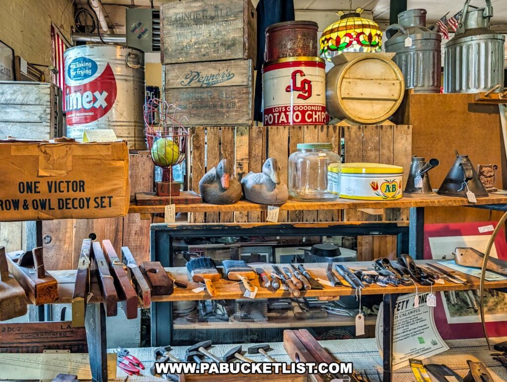 Rustic display at the Penn Avenue Trading Company in Newport, Perry County, PA, featuring an assortment of vintage hand tools, wooden planes, decoys, advertising tins, glass jars, oil cans, and wooden crates, all arranged on weathered shelves beneath a stained-glass lamp in this multi-vendor antique store.