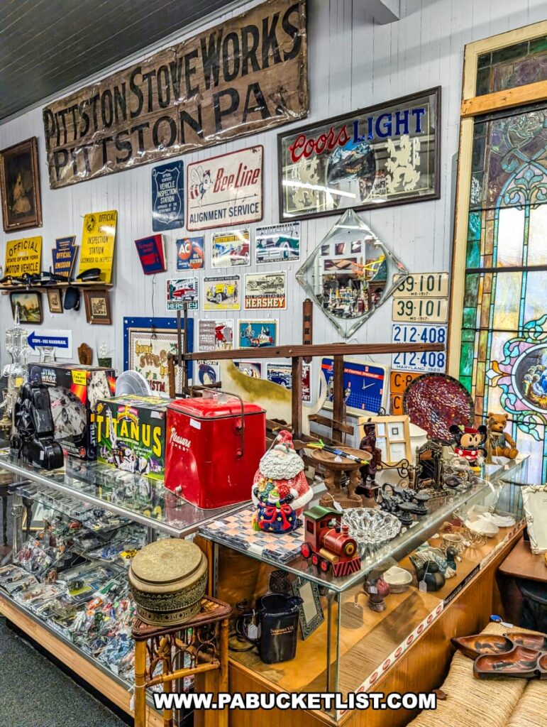 A vibrant interior view of Duchess Outlet Antiques, a multi-vendor store housed in the old Pittston Stove Building in Pittston, PA. The image showcases a diverse array of antiques, collectibles, and vintage goods displayed across multiple shelves and a large glass counter, with prominent items including old advertising signs like "PITTSTON STOVE WORKS," "Coors LIGHT," and "BeeLine," along with a red vintage cooler, Christmas decor, and other unique finds under glass. A stained glass window is visible on the right, adding to the historic ambiance.