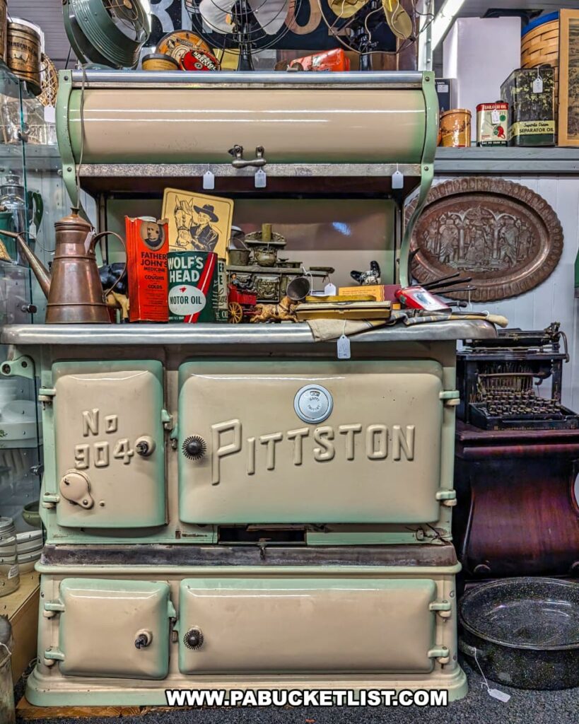 An antique Pittston stove, cream and pale green, prominently displayed in a multi-vendor antique store, surrounded by vintage collectibles like oil cans, a copper kettle, and a typewriter, with other antique items visible in the background, showcasing the diverse offerings of an antique shop housed in the historic Pittston Stove Building.