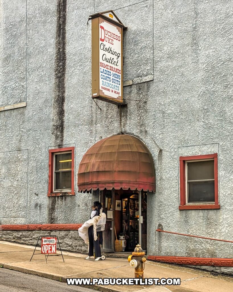 Exterior view of Duchess Outlet Antiques in Luzerne County, PA, showing the old Pittston Stove Building with a red awning entrance, mannequin display, open antiques sign on the sidewalk, and a vintage wall sign advertising clothing and outerwear.