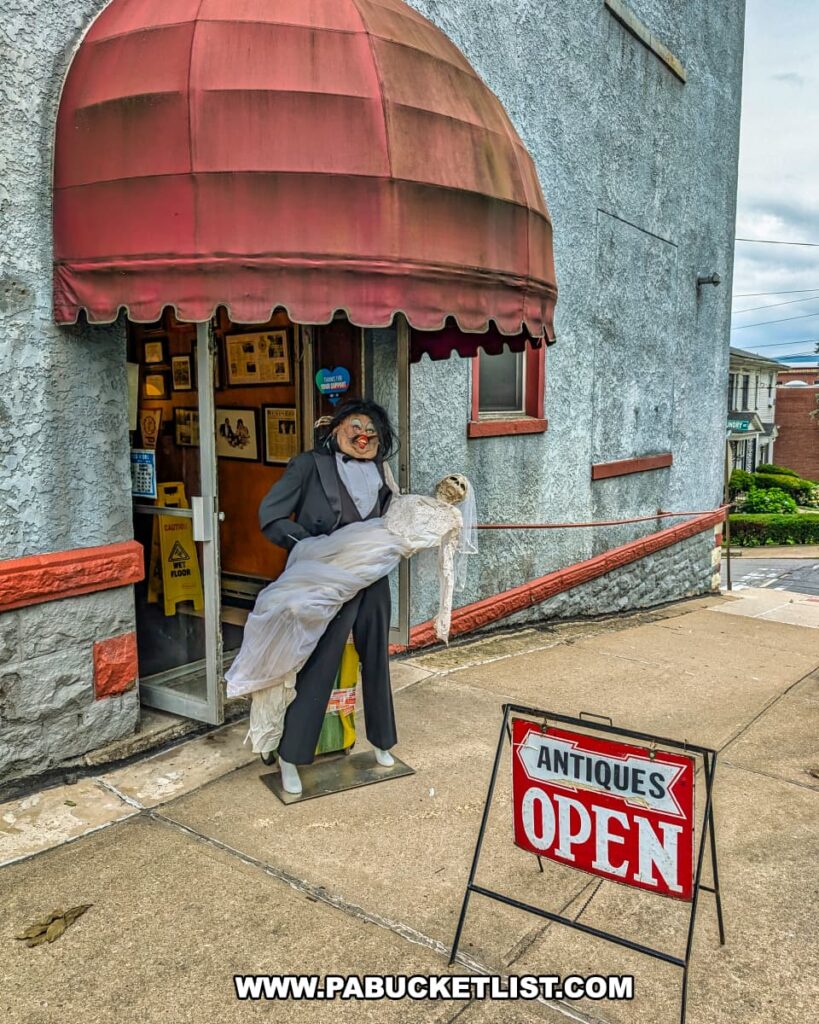 Front entrance of Duchess Outlet Antiques in Pittston, PA, featuring a mannequin display of a tuxedo-clad figure carrying a doll beneath a red awning, with an “Antiques Open” sign placed on the sidewalk.