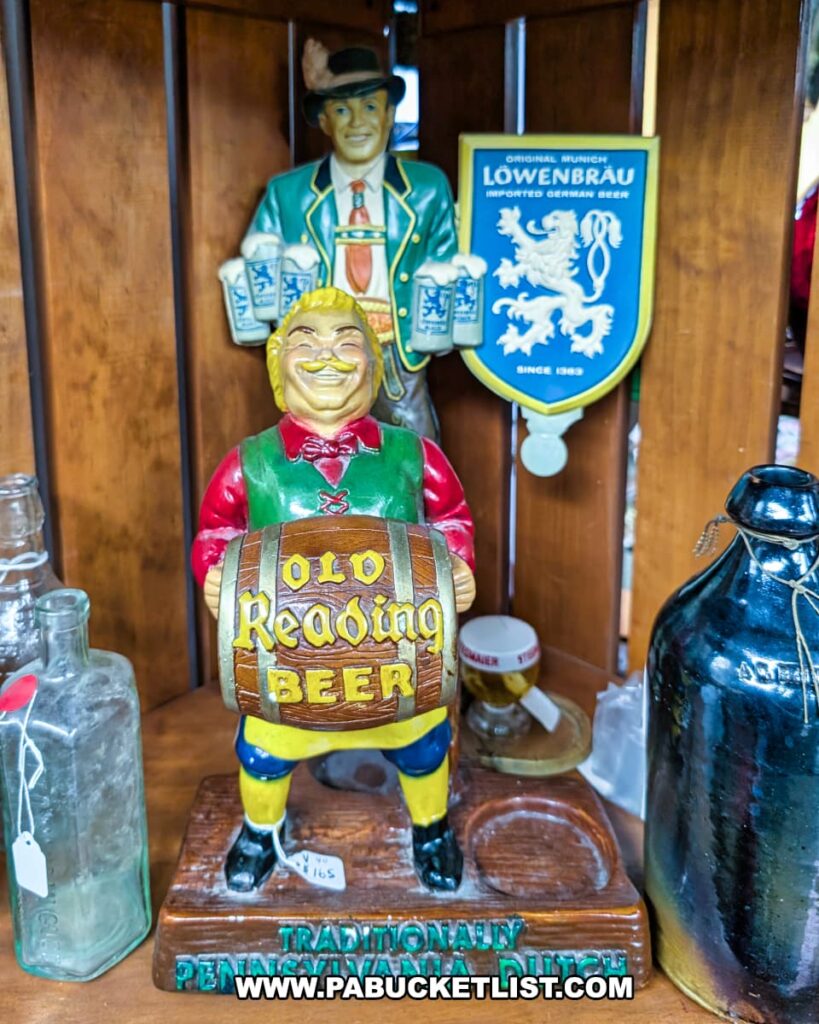 A close-up view inside an antique store display case showcases two vintage beer advertising figures: a smiling, plump man in a green vest and red shirt holding a beer barrel labeled "Old Reading Beer," positioned in front of a taller figure of a man in traditional Bavarian attire standing beside a shield-shaped sign for "Löwenbräu Imported German Beer." Various antique glass bottles and jugs are visible on the shelves around the figures, suggesting a collection of vintage barware and advertising memorabilia.