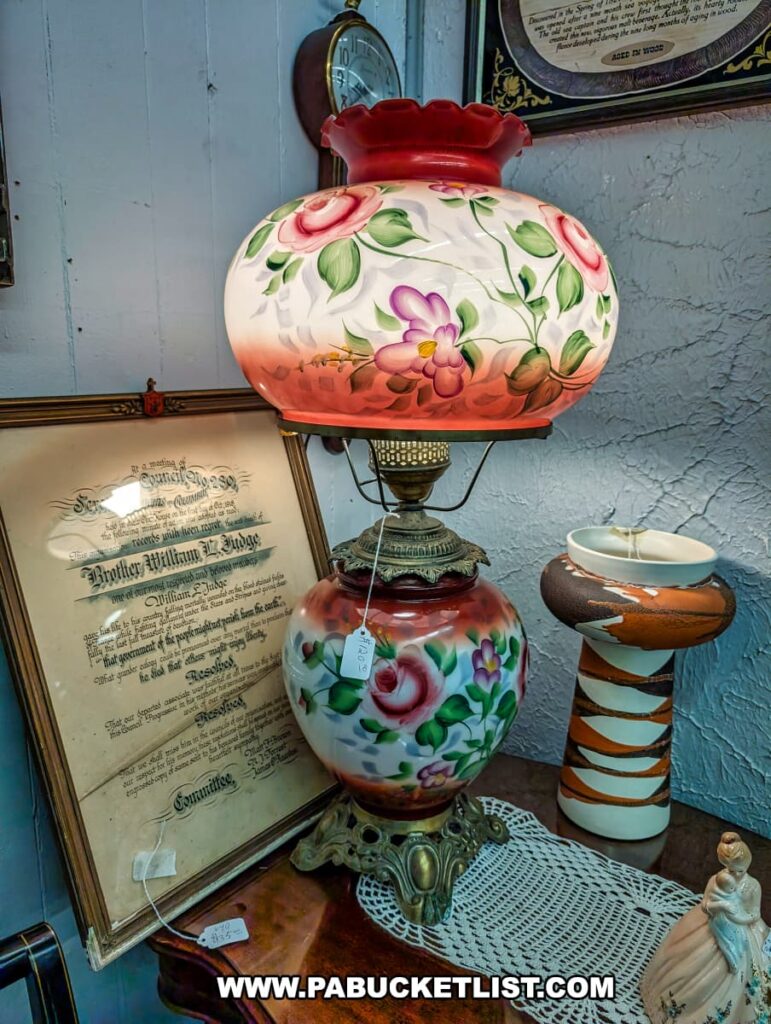 A vintage "Gone with the Wind" style lamp with hand-painted floral designs in shades of red, pink, and green sits on a dark wooden table covered with a white doily.