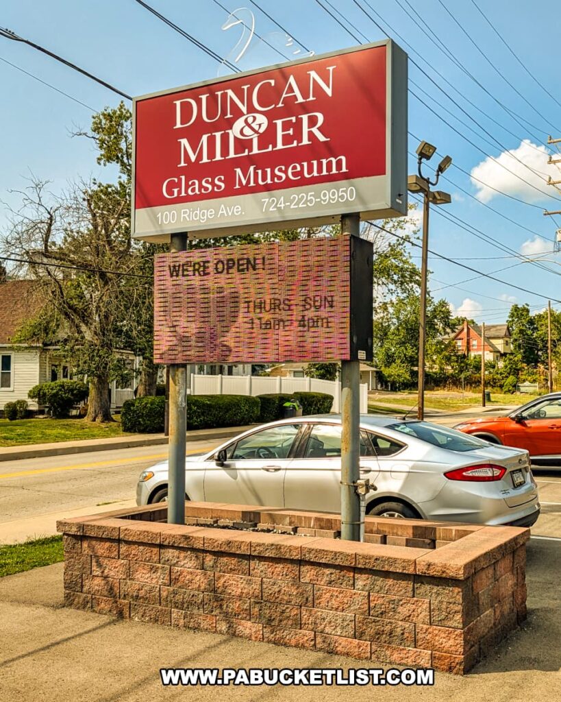 Entrance sign for the Duncan and Miller Glass Museum in Washington, PA, displaying the museum’s name, address, phone number, and open hours against a sunny backdrop.