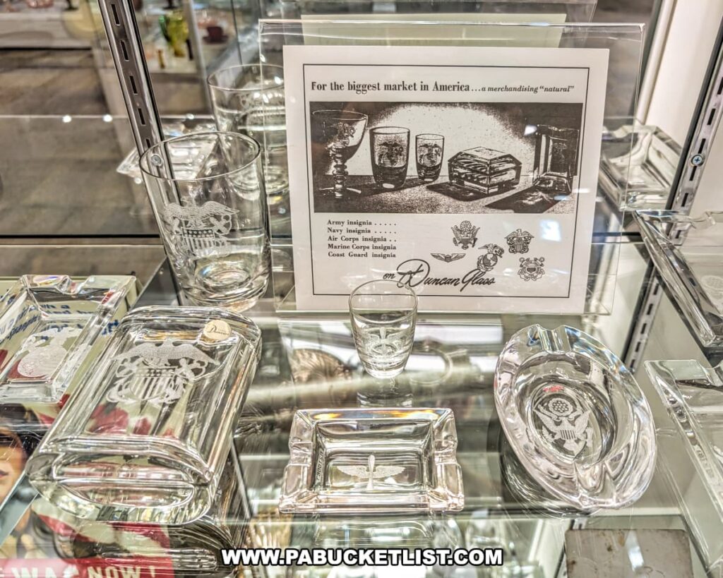 Display of military-themed glassware at the Duncan and Miller Glass Museum in Washington, PA, featuring etched insignias for the Army, Navy, Air Corps, Marines, and Coast Guard on glasses, trays, and ashtrays produced during World War II.