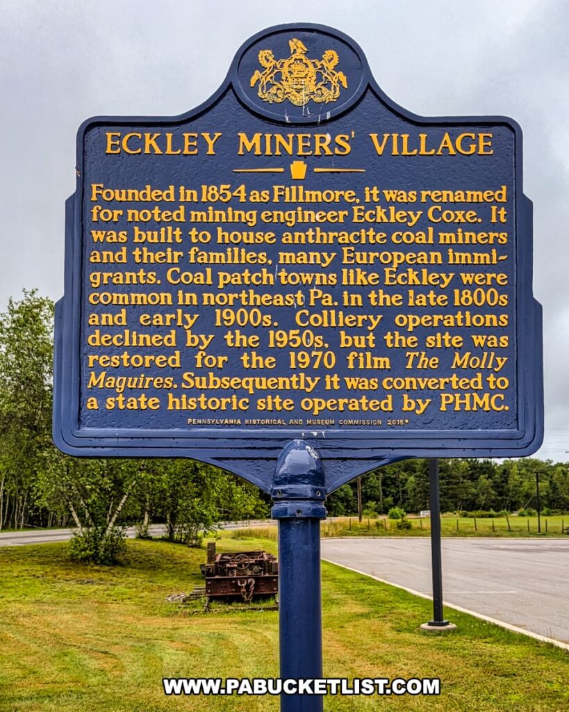 State historical marker at Eckley Miners’ Village in Luzerne County, Pennsylvania, detailing the town’s founding in 1854, its role as a coal patch community for anthracite miners and European immigrants, its decline by the 1950s, restoration for the 1970 film *The Molly Maguires*, and preservation as a state historic site operated by the Pennsylvania Historical and Museum Commission.