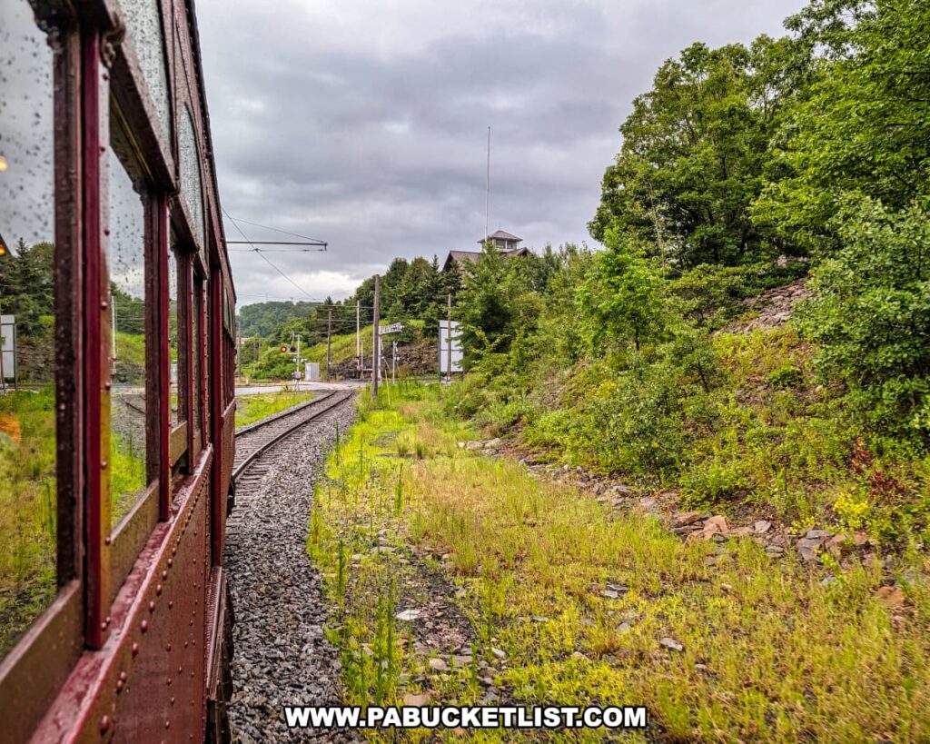 View from a vintage trolley car on the Electric City Trolley Museum’s 5.5 mile round-trip excursion in Lackawanna County, showing the track curving through a green, wooded hillside under cloudy skies.