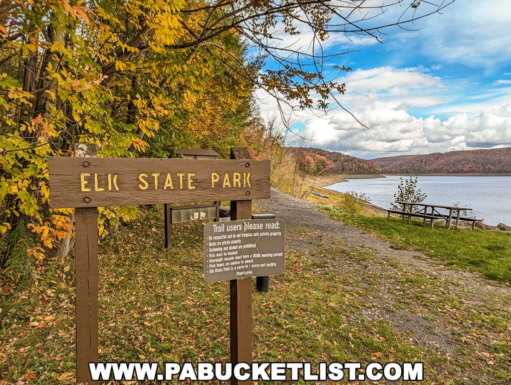 Wooden Elk State Park sign surrounded by brilliant yellow and orange autumn leaves, with a lake, picnic tables, and colorful hillsides under a partly cloudy sky in the background.
