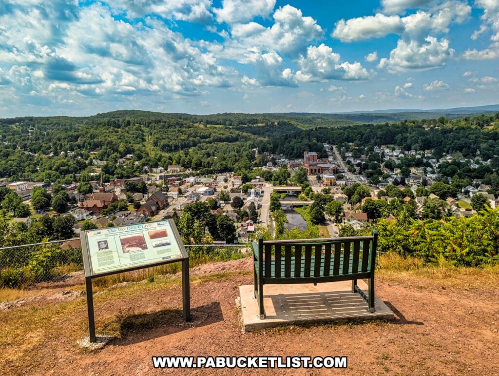 Bench and informational sign at Irving Cliff overlook in Wayne County, PA, offering a sweeping summer view of the town of Honesdale nestled among forested hills under a partly cloudy sky.