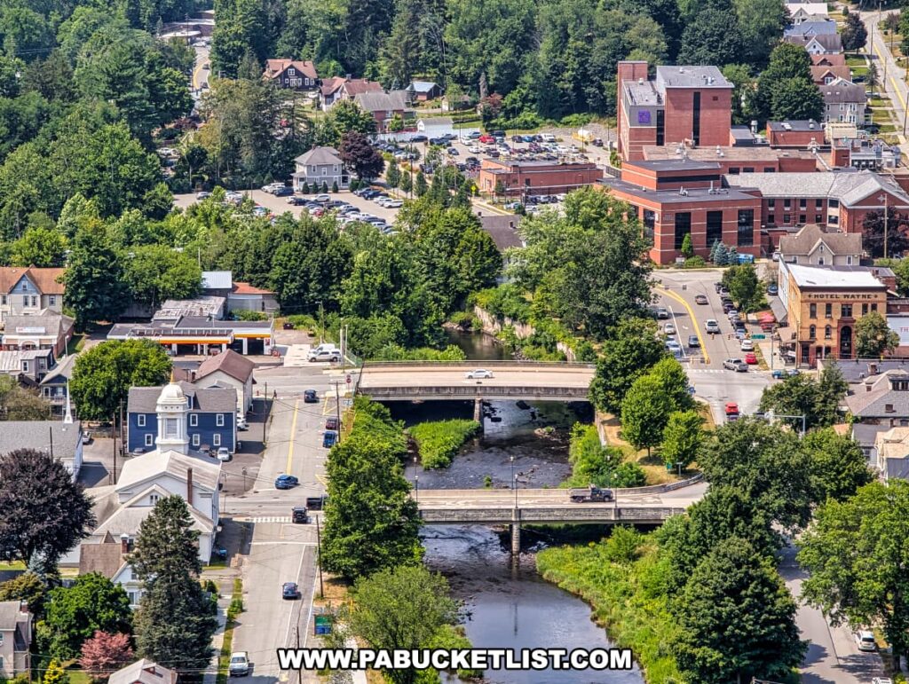 Aerial view from Irving Cliff looking down into downtown Honesdale, Pennsylvania, highlighting two bridges crossing the Lackawaxen River, the historic Hotel Wayne, a church with a white steeple, and several red brick buildings surrounded by tree-lined streets and a mix of homes and businesses.