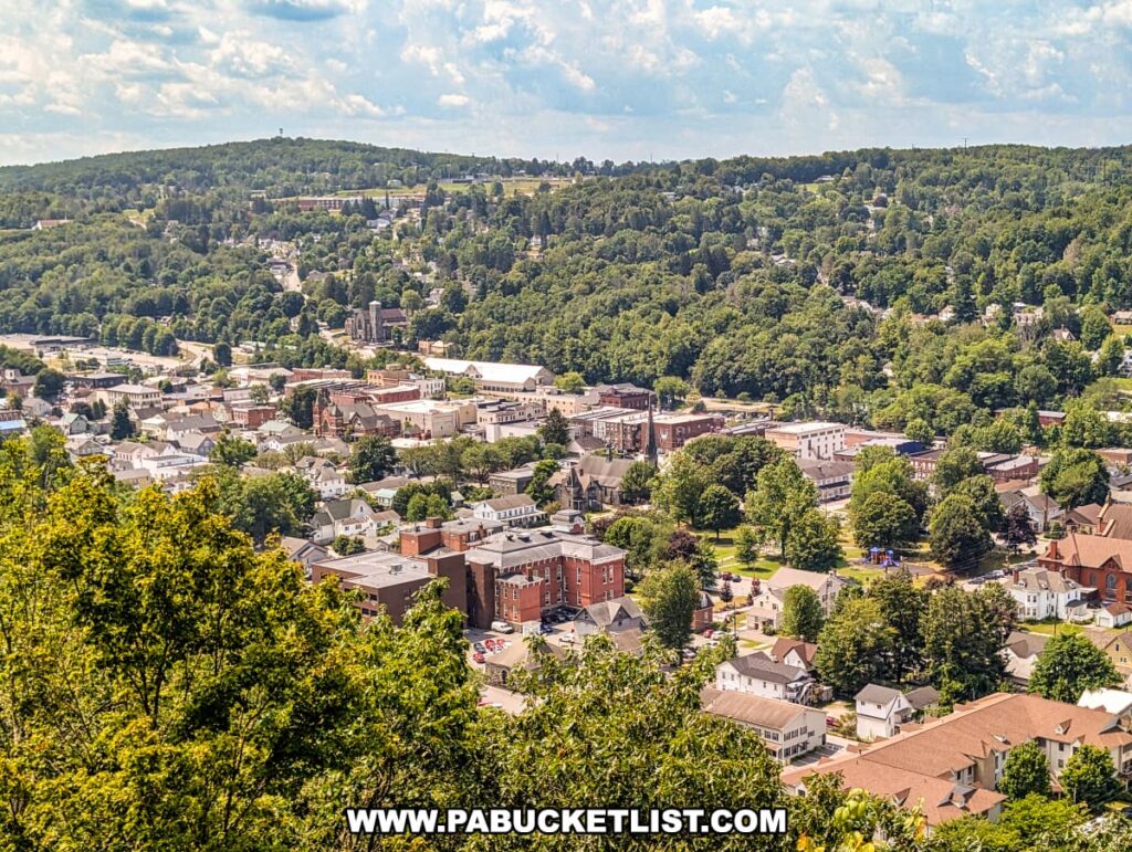 Expansive view from Irving Cliff in Wayne County, PA, looking south over the town of Honesdale, with tree-lined neighborhoods, historic brick buildings, church steeples, and rolling forested hills under a partly cloudy summer sky.