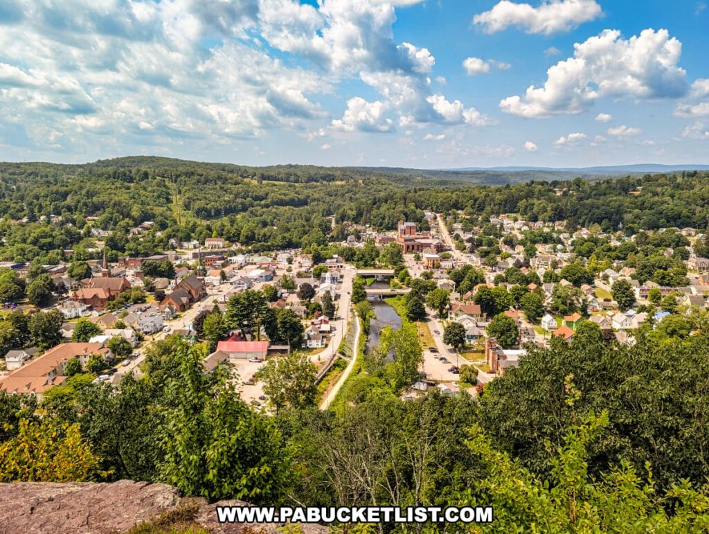 Sweeping summer view from Irving Cliff in Wayne County, PA, looking west over the town of Honesdale, with the Lackawaxen River winding through the heart of town, framed by historic churches, downtown buildings, tree-lined streets, and rolling green hills in the distance.