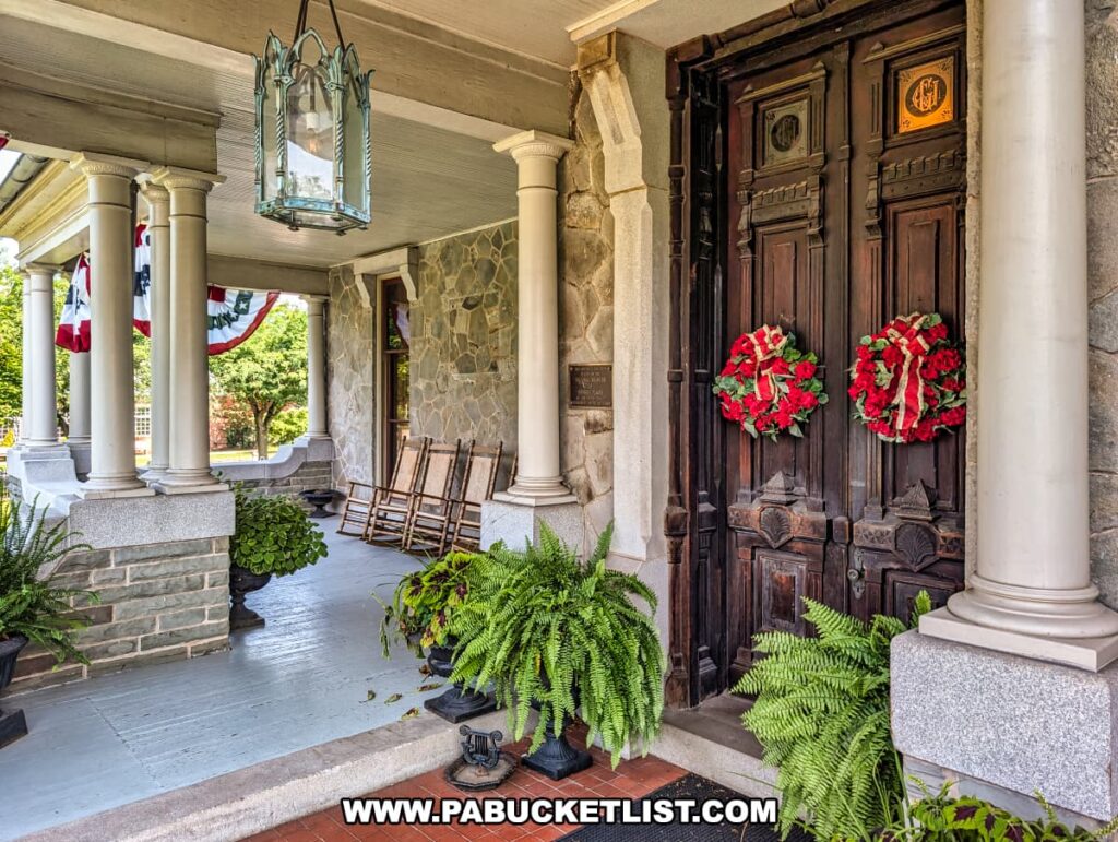 Front porch of the Jackson Mansion featuring tall columns, hanging lanterns, potted ferns, wooden rocking chairs, patriotic bunting, and ornate double doors adorned with red wreaths.