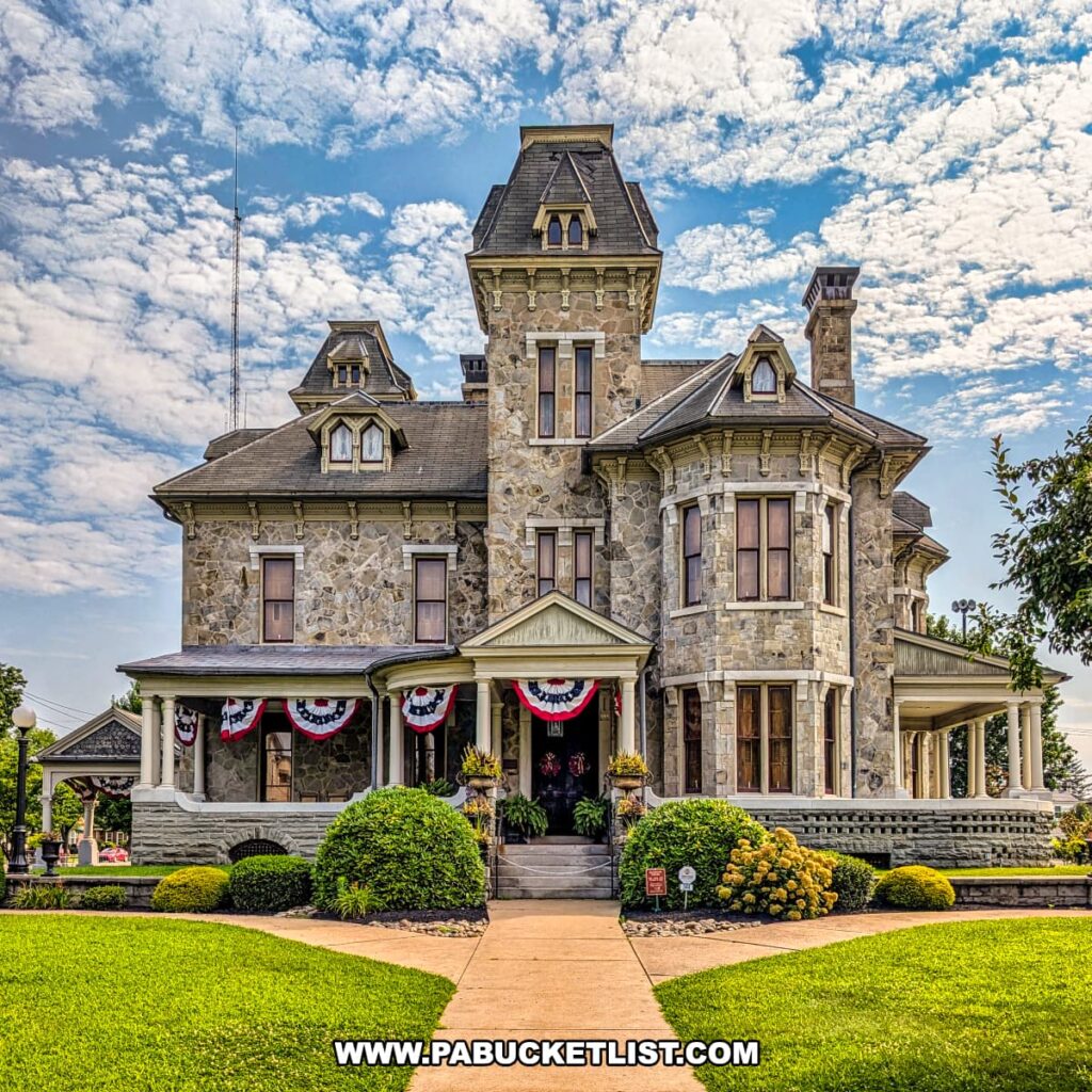 Front view of the Jackson Mansion in Berwick, Pennsylvania, showcasing its grand Second Empire/Italianate architecture, central tower, wraparound porch with patriotic bunting, and landscaped gardens beneath a partly cloudy sky.