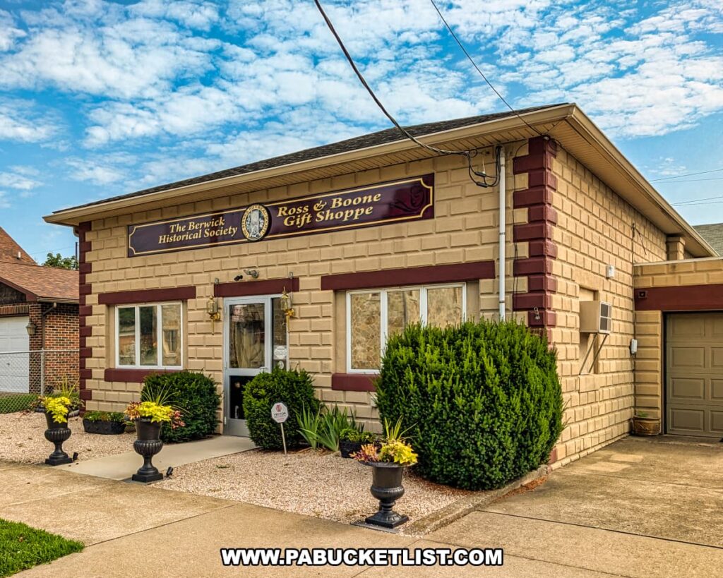 Ross & Boone Gift Shoppe, operated by the Berwick Historical Society near the Jackson Mansion, featuring a tan brick exterior with burgundy trim, decorative planters, and a welcoming entrance under a partly cloudy sky.
