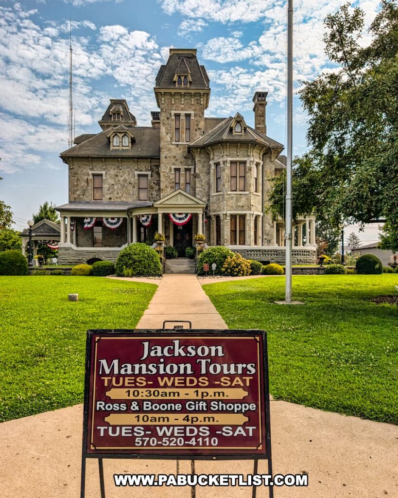 Front view of the Jackson Mansion in Berwick, Pennsylvania, with a tour hours sign for the Berwick Historical Society displayed along the walkway leading to the grand stone home adorned with patriotic bunting.