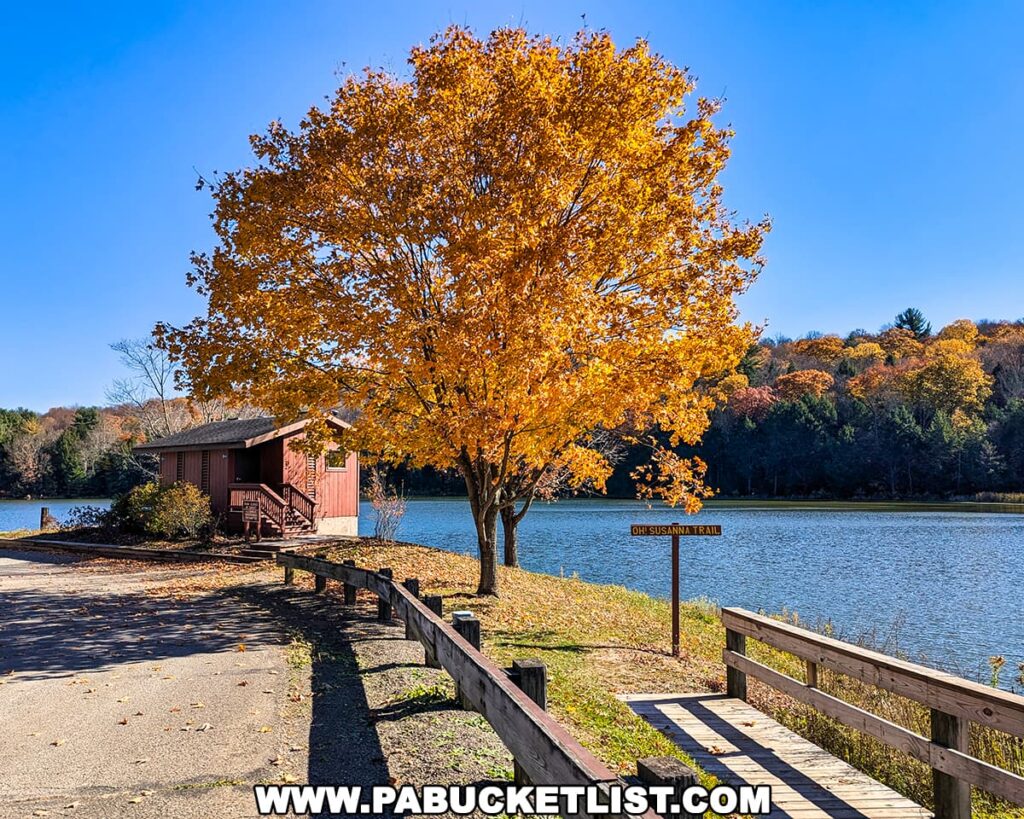 Brilliant orange fall foliage frames the Oh! Susanna Trail sign and a rustic lakeside building at Mount Pisgah State Park, with wooded hillsides glowing in autumn colors under a clear blue sky.