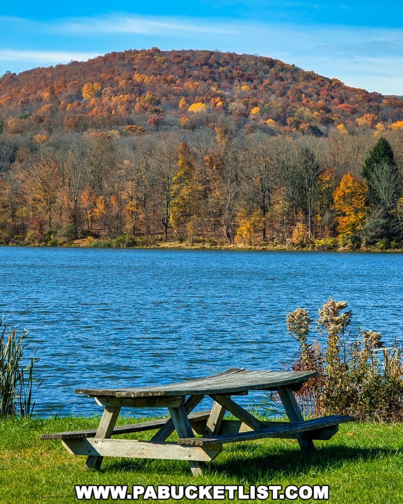 Wooden picnic table by the lake at Mount Pisgah State Park with hillsides ablaze in vibrant shades of red, orange, and gold autumn foliage under a clear blue sky.