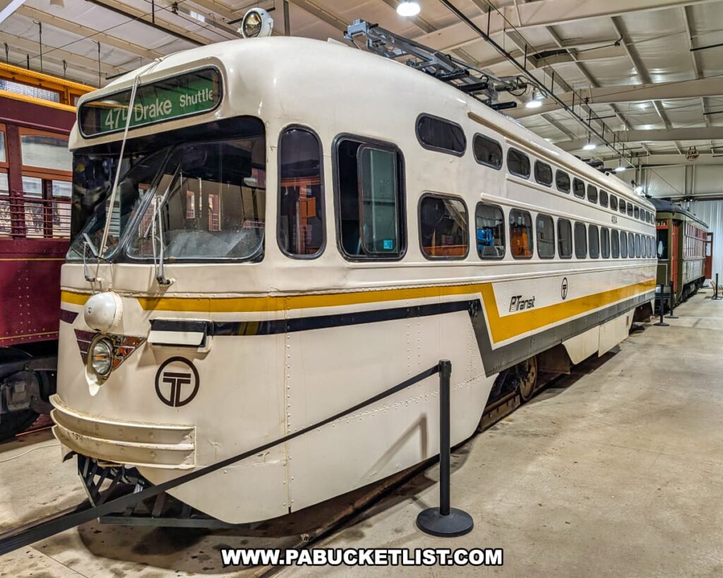 Vintage PCC streetcar painted in white with gold and black stripes on display inside the Pennsylvania Trolley Museum in Washington County, Pennsylvania, featuring a 47D Drake Shuttle route sign and surrounded by other historic trolleys.