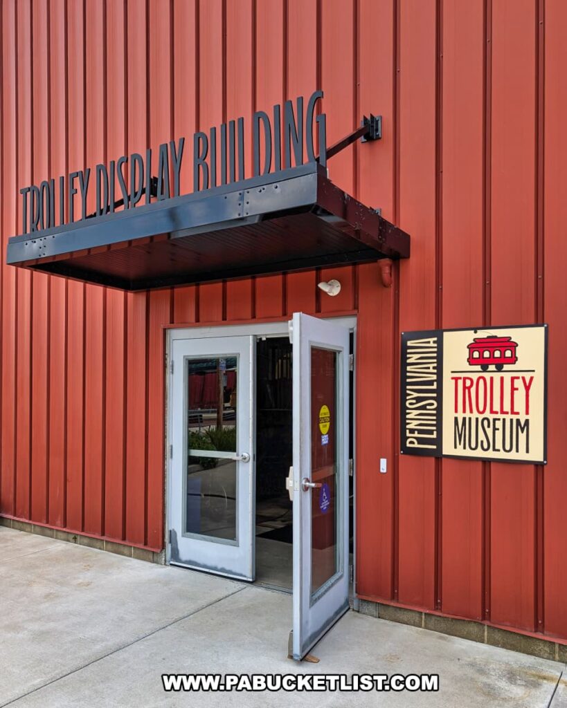 Entrance to the Trolley Display Building at the Pennsylvania Trolley Museum in Washington County, Pennsylvania, featuring a red exterior wall, double glass doors, and a sign with a vintage trolley graphic welcoming visitors.
