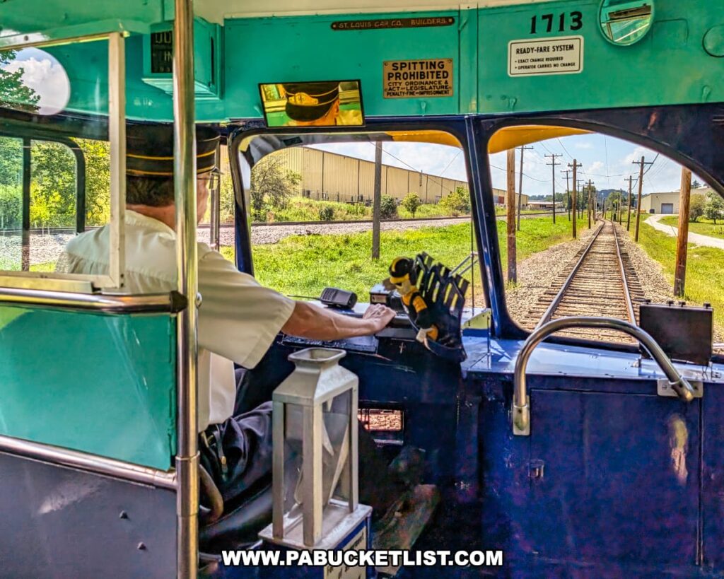 Motorman operating a vintage trolley at the Pennsylvania Trolley Museum in Washington County, Pennsylvania, with a view through the front windows of the trolley tracks stretching into the distance on a sunny day.
