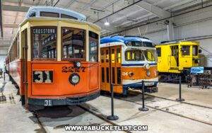 Trio of vintage trolleys on display inside the Trolley Display Building at the Pennsylvania Trolley Museum in Washington County, Pennsylvania, including an orange Crosstown car, a blue and orange Media car, and a yellow maintenance trolley.