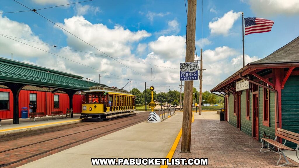 Yellow open-air vintage trolley arriving at the boarding platform behind the Pennsylvania Trolley Museum in Washington County, Pennsylvania, with the historic Wexford Station building and an American flag visible nearby.