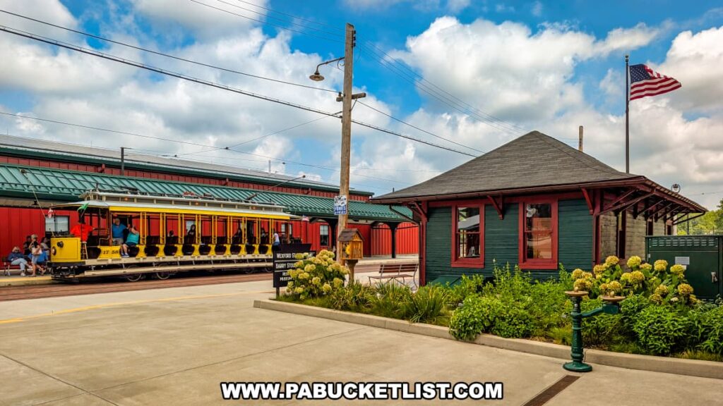 Open-air yellow and red vintage trolley filled with passengers at the boarding platform behind the Pennsylvania Trolley Museum in Washington County, Pennsylvania, with the historic Wexford Station and American flag in the background.