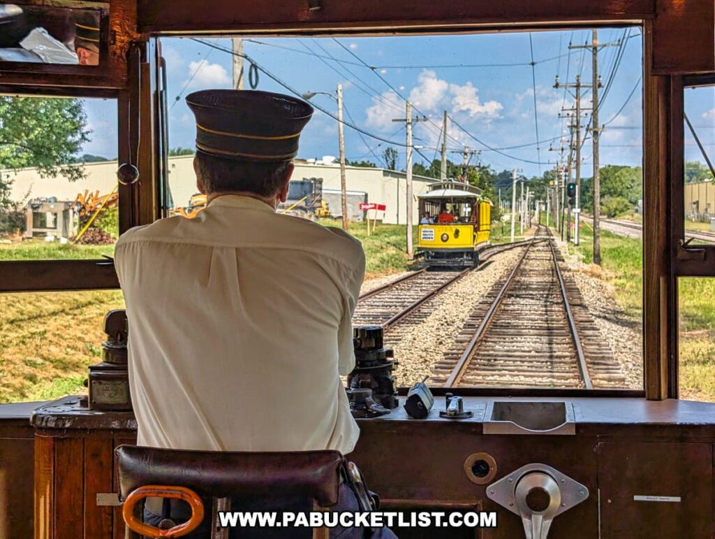 View from inside a vintage trolley at the Pennsylvania Trolley Museum in Washington County, Pennsylvania, showing the motorman operating the controls as another yellow open-air trolley passes on the adjacent track during the scenic excursion ride.