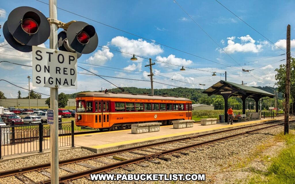 Bright orange vintage Valley Route trolley stopped at the Washington County Fairgrounds platform outside the Pennsylvania Trolley Museum in Washington County, Pennsylvania, with a railroad signal and surrounding hills visible in the background.