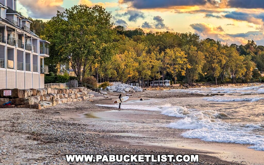Surfer standing at the edge of Lake Erie with waves crashing on the shore at Presque Isle State Park, backed by golden autumn foliage and a dramatic sunset sky.