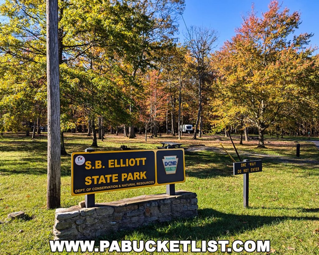 Entrance sign for S.B. Elliott State Park surrounded by a mix of golden yellow, orange, and red autumn foliage under a bright blue sky.