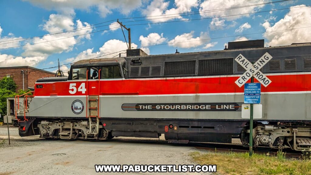 Stourbridge Line locomotive number 54, painted in red, white, and black, crosses a road in Honesdale, Wayne County, Pennsylvania, beneath a bright blue sky with scattered clouds and a visible railroad crossing sign in the foreground.