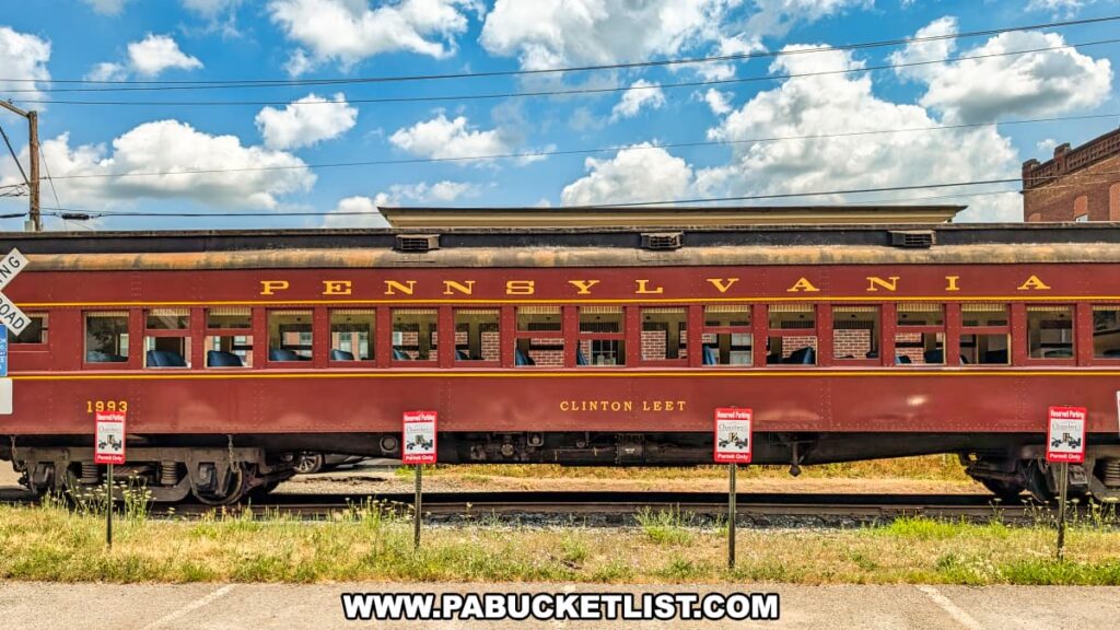 A maroon and gold vintage passenger car labeled "PENNSYLVANIA" and named "Clinton Leet" sits on the tracks in Honesdale, Wayne County, Pennsylvania, as part of the Stourbridge Line sightseeing excursion, with a clear blue sky and historic brick buildings in the background.