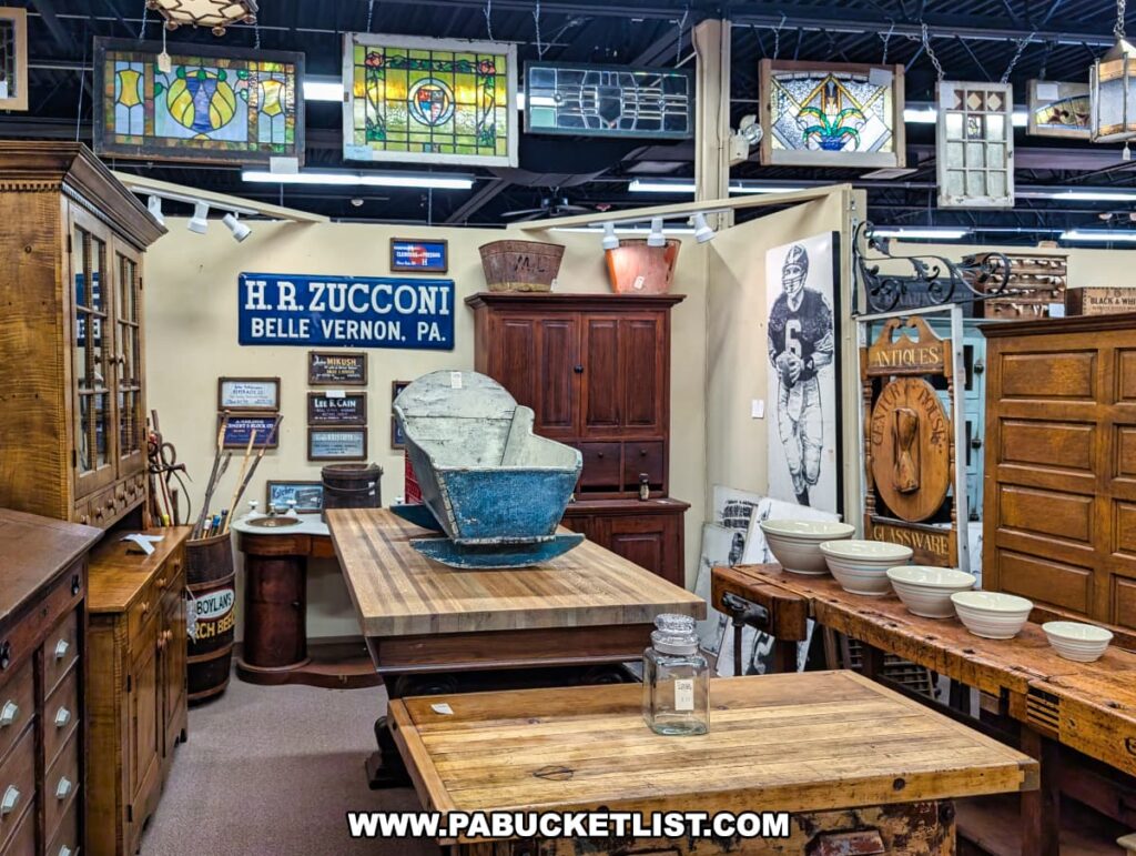 Display of antique furniture and decorative arts at the Antique Center of Strabane in Canonsburg, Pennsylvania, featuring wooden cabinets, farmhouse tables, stoneware bowls, vintage advertising signs, stained glass windows, and a large blue wooden dough trough centerpiece.