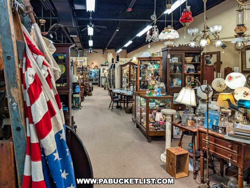Aisle at the Antique Center of Strabane in Canonsburg, Pennsylvania, lined with vendor booths displaying 18th through early 20th century antiques including glassware, lamps, chandeliers, furniture, and Americana décor such as a draped American flag.