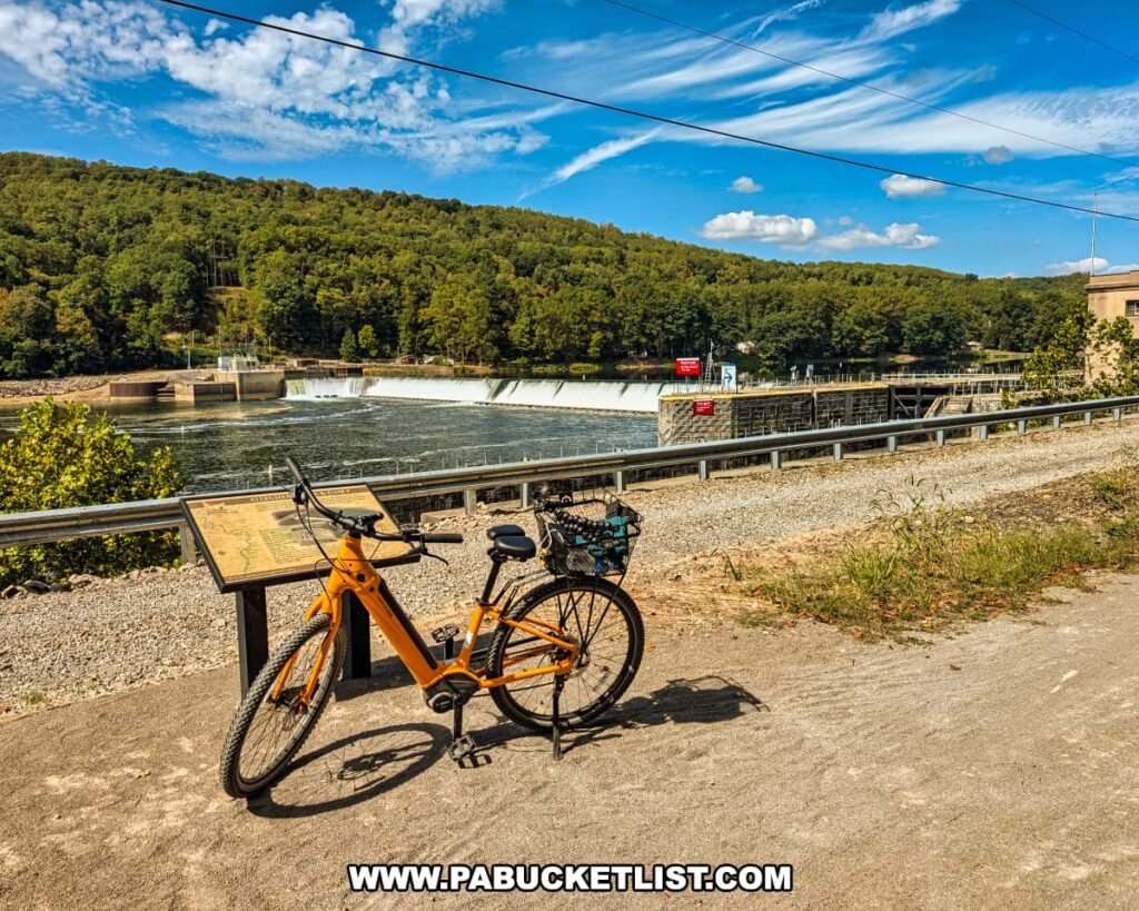 Yellow bicycle parked along the Armstrong Trail in western Pennsylvania, with an informational sign in the foreground and Lock and Dam Number 9 spanning the Allegheny River against a backdrop of forested hills and a bright blue sky.