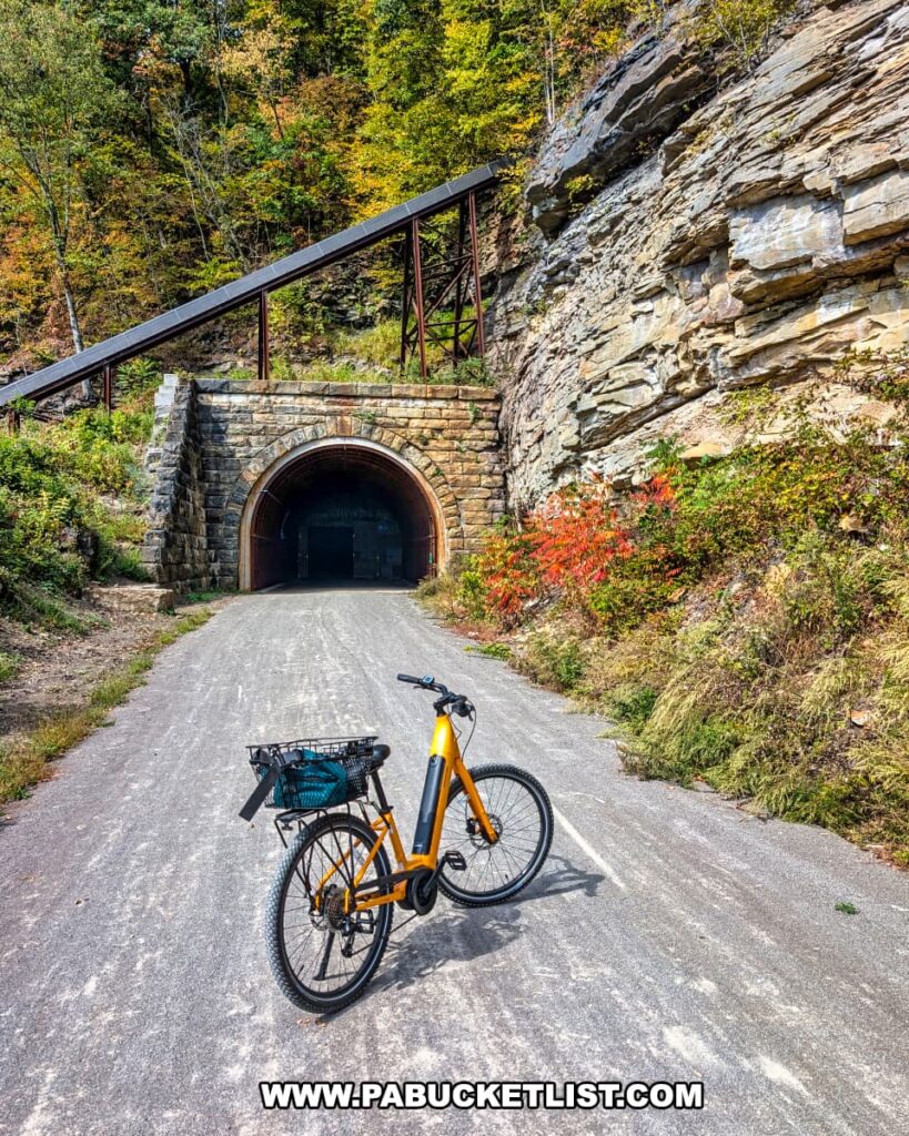 Yellow bicycle parked in front of the stone entrance to the Brady Tunnel along the Armstrong Trail in western Pennsylvania, with colorful autumn foliage and exposed rock cliffs surrounding the tunnel portal.