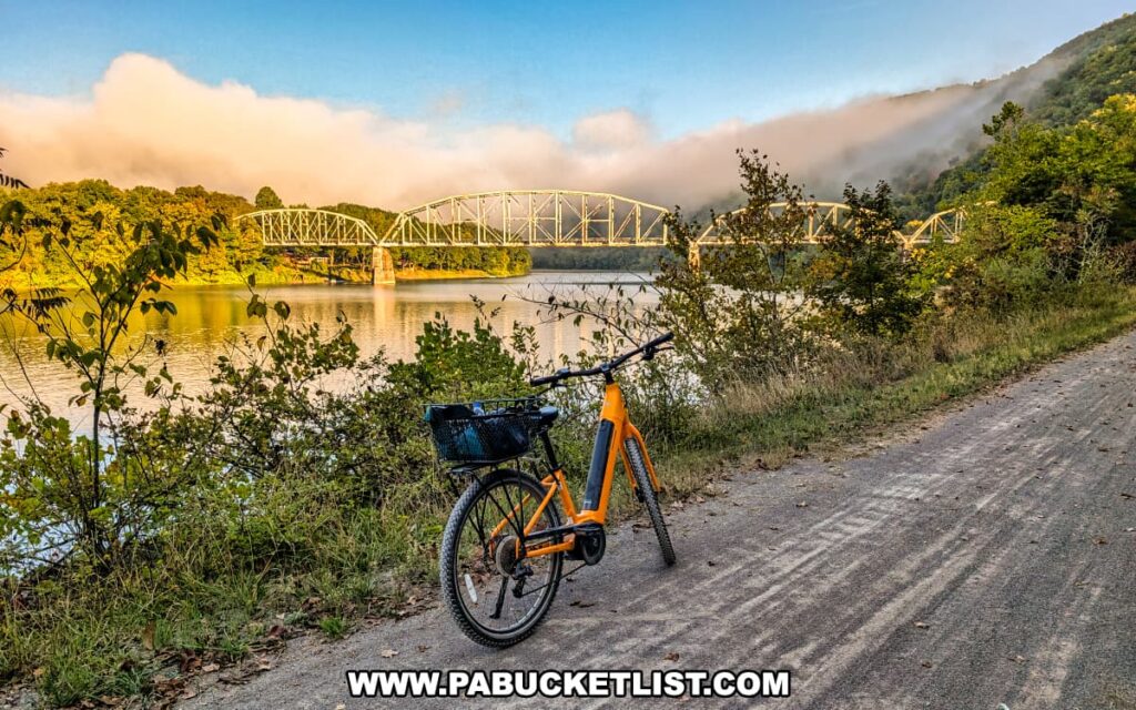 Yellow bicycle parked along the Armstrong Trail in western Pennsylvania, with the Pittsburgh and Shawmut Railroad Bridge spanning the Allegheny River in the background, framed by morning fog and sunlit hillsides.