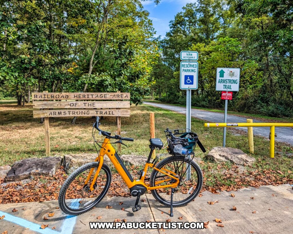 Yellow bicycle parked at the northern trailhead of the Armstrong Trail in western Pennsylvania, next to signs marking the Railroad Heritage Area and trail access, with a tree-lined path extending into the distance.
